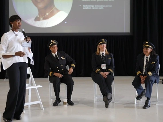 A Black woman in a white dress shirt and black pants stands with a microphone in front of three people in airline pilot uniforms who are seated in white chairs on a stage in front of a black background.