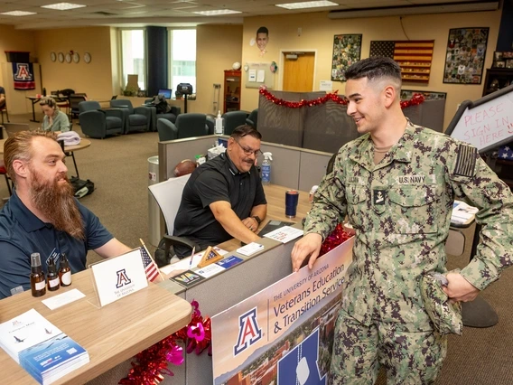 A U.S. Navy service member in a camouflage uniform chats with two men seated at a front desk in a military student support center. The desk is decorated with American flags, brochures, and a sign reading "Veterans Education & Transition Services" from the University of Arizona. The setting is an inviting space with couches, decorations, and a whiteboard that says, "Please sign in here."