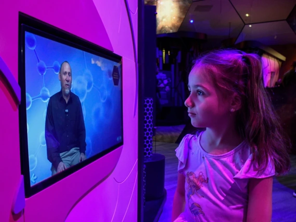 A young girl with long brown hair and a pink mermaid shirt gazes at an interactive screen in a science exhibit. The screen displays a man speaking against a blue molecular-patterned background. The exhibit is illuminated with vibrant purple and blue lighting.