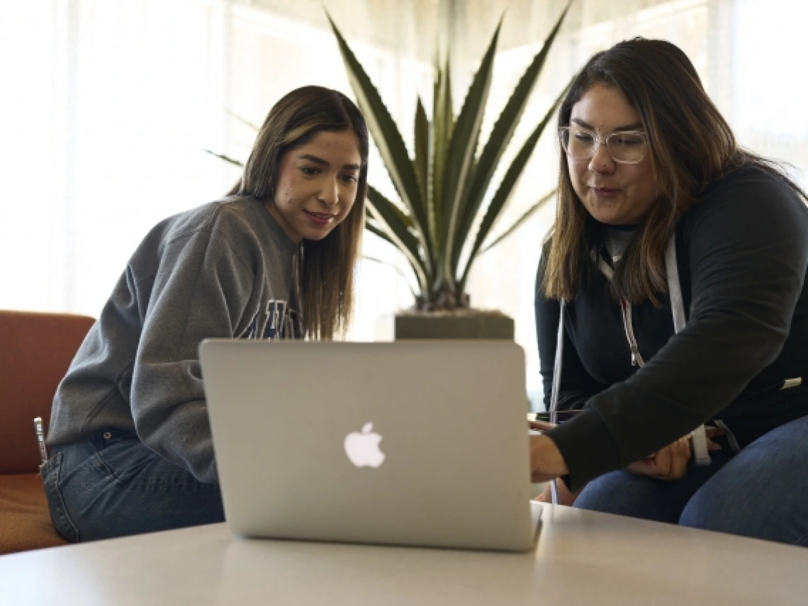 Two students sitting at a table looking at a laptop computer.